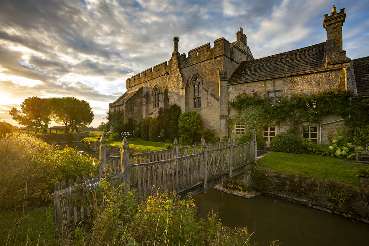 A photograph of Markenfield Hall taken at 6:00am, when the sun was just rising behind the trees. In the foreground are some wild-growing native plants, and then the wooden footbridge that leads over the moat and to the Hall itself. The Hall is illuminated by the first rays of the sun, which glint on the small glass panes of the medieval arched windows. The blue sky is streaked with clouds. The whole photograp is bathed in a golden light.